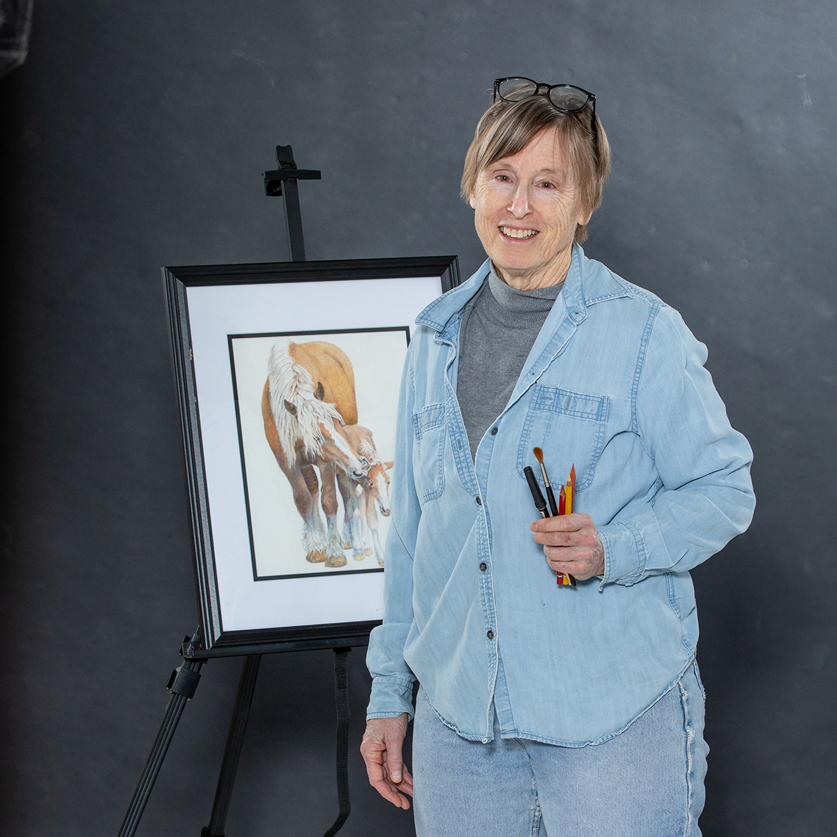 Marcia smiling beside an easel holding a framed watercolor of Haflinger draft horses, paintbrushes and pencils in hand